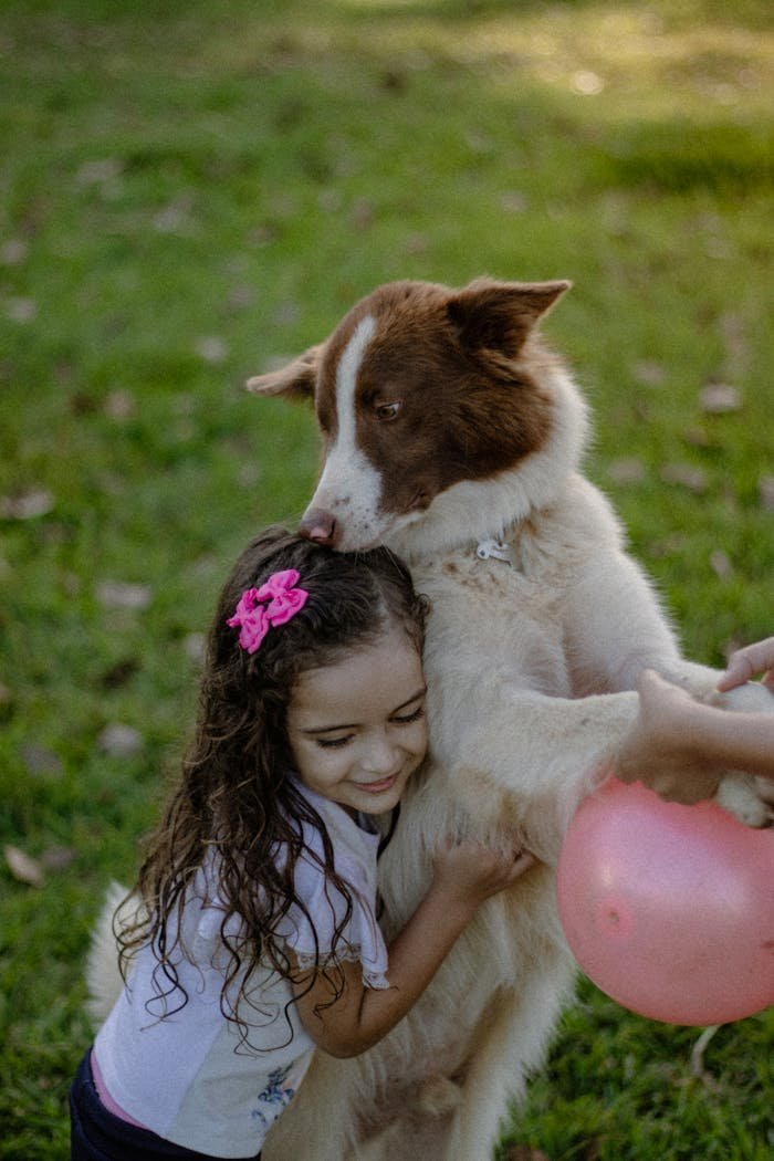 A joyful child hugging a dog while holding a pink balloon on a sunny day outdoors.